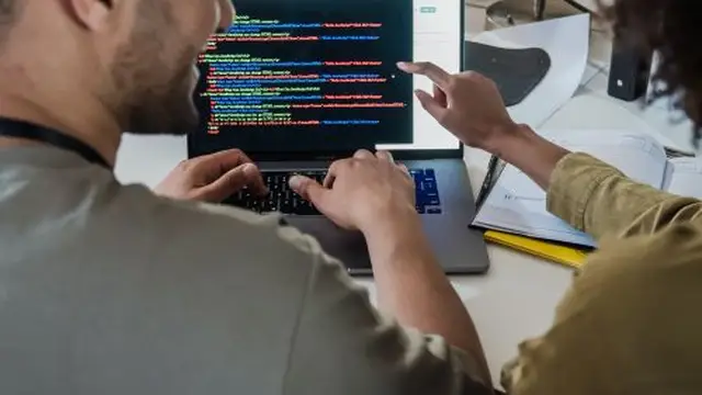 Two developers working side by side on code at a desk with a laptop and monitors