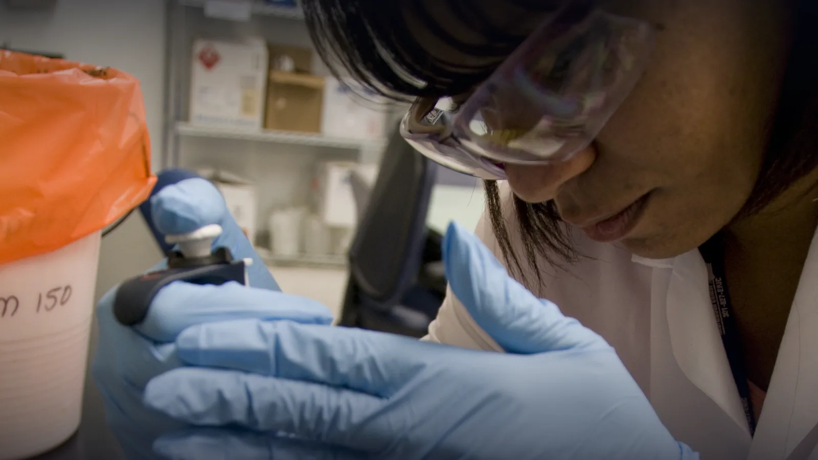A microbiologist in a laboratory handling a pipette while wearing gloves and safety glasses