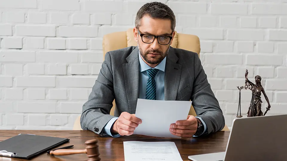 A lawyer at a desk reviewing a document beside a laptop, gavel and Lady Justice statuette