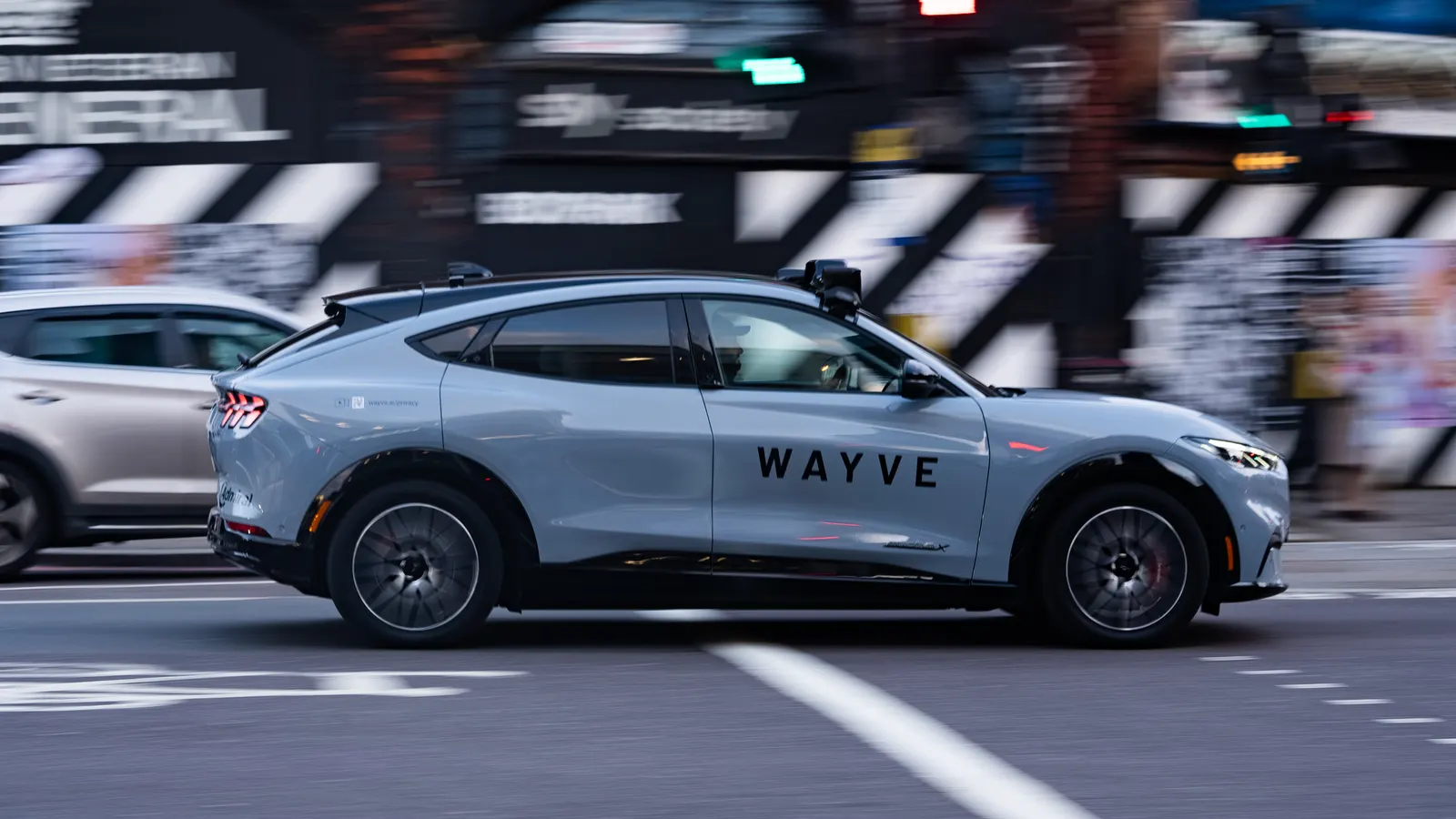 A Wayve autonomous test vehicle, a Ford Mustang Mach-E with Wayve livery and roof-mounted sensors, driving on a London street